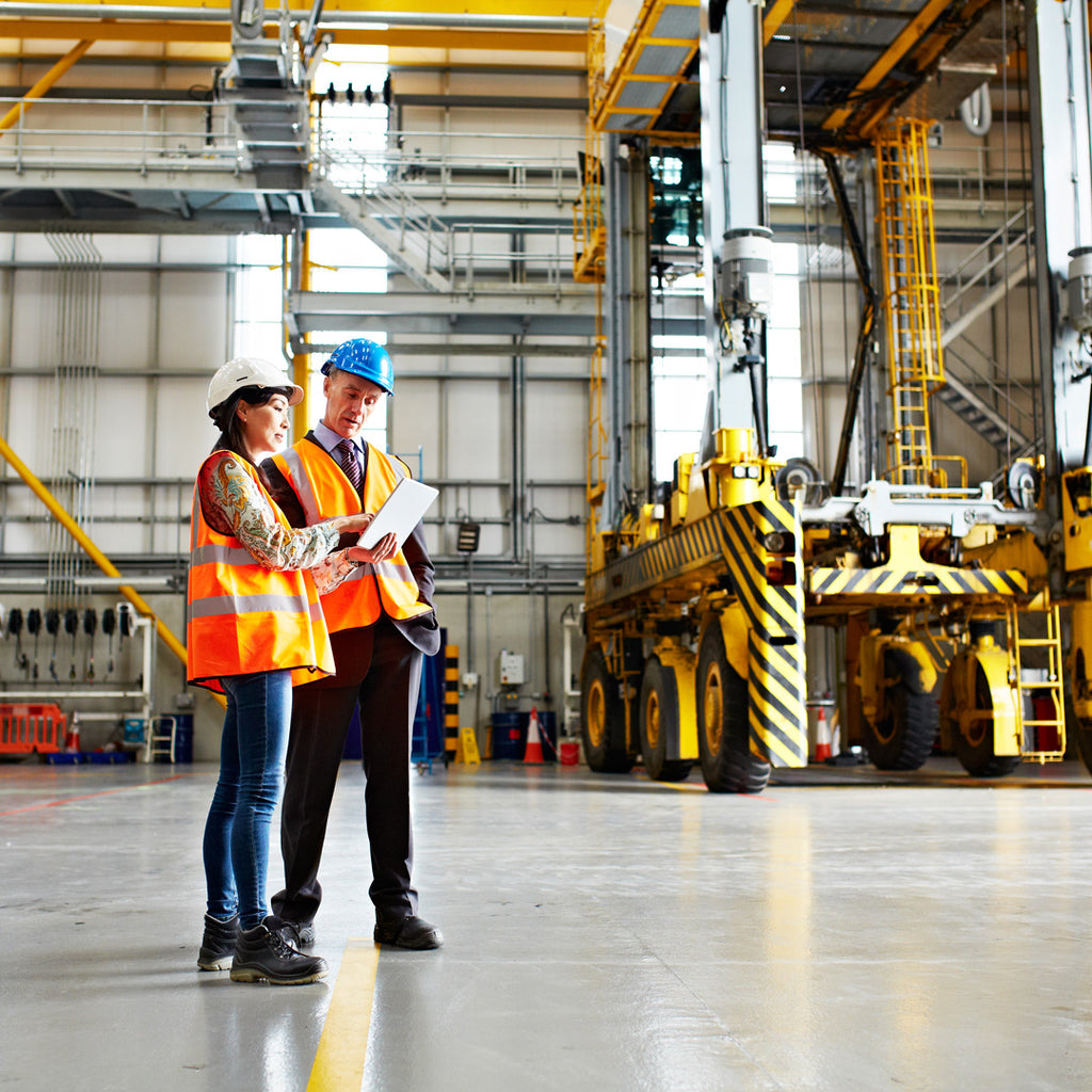 Two professionals discussing safety protocols in a well-lit warehouse, with heavy machinery in the background, symbolizing the importance of safety measures like strip curtains in industrial environments.
