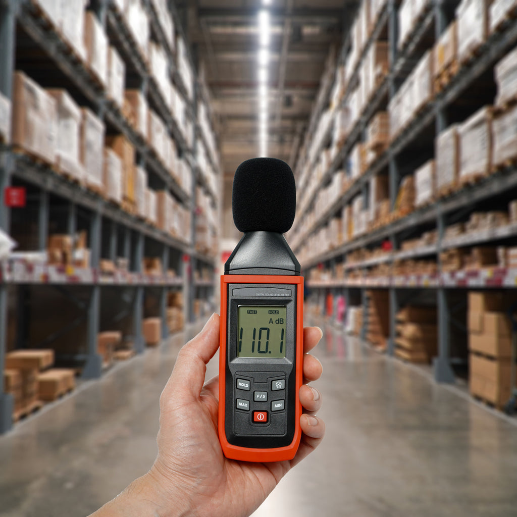 Hand holding a decibel meter showing 101.0 dB in a warehouse, illustrating noise levels and the need for effective noise reduction strategies.