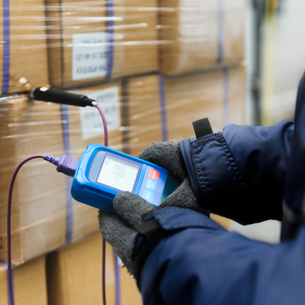 Close-up of a gloved hand holding a digital thermometer, measuring temperature in a warehouse, highlighting the effectiveness of strip curtains in maintaining optimal conditions.