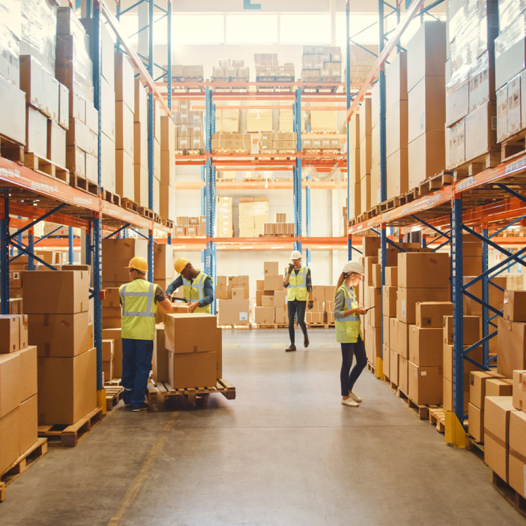 Warehouse aisle with pallet racking and boxed stock; workers in hi-vis managing inventory and picking in a busy storage area.