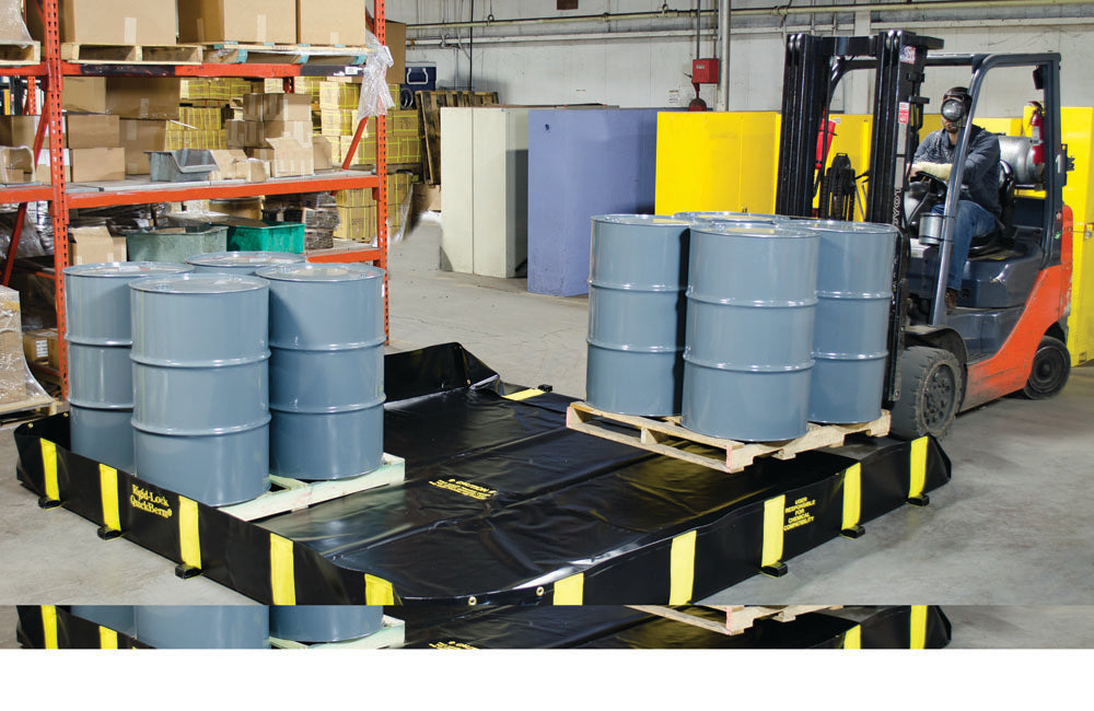 A black and yellow rigid-lock quick berm used for spill containment, shown with several gray drums on top, placed in an industrial setting with a forklift in the background.